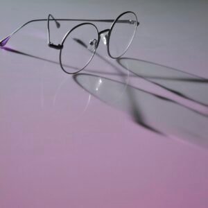Round eyeglasses with metal frames casting shadows on a neon purple surface, emphasizing modern eyewear style.